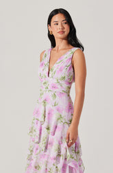 A woman is wearing a sleeveless, V-neck dress with a fitted bodice that features a gathered design and a waistline accentuated with a wide band. The dress has a floral pattern with pink and green flowers on a white background. The skirt is tiered with ruffled layers, creating a flowing and layered appearance.